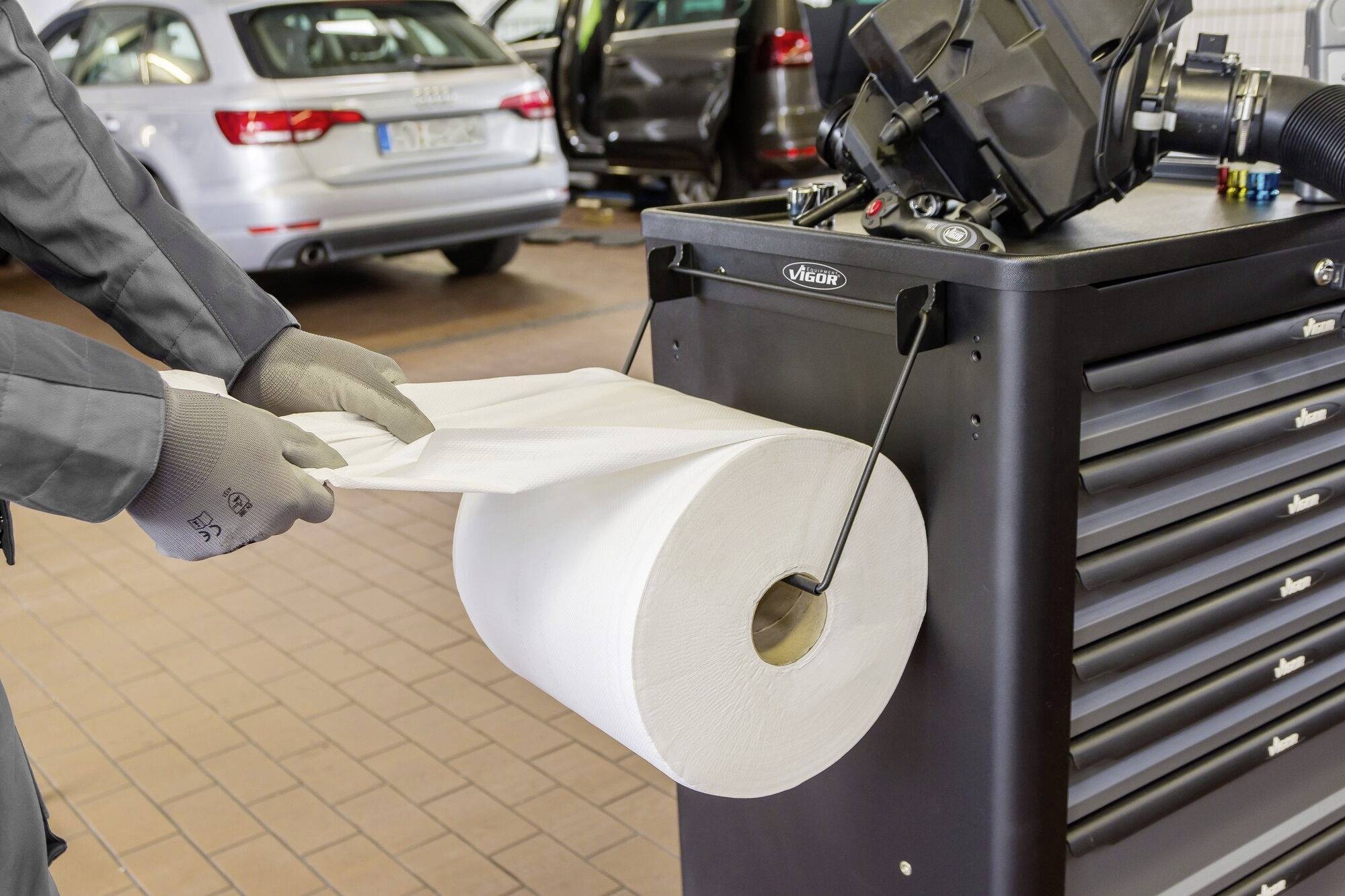 Een persoon met handschoenen trekt een papieren doek van een grote rol naast een werkbank in een autogarage. Autobanden zijn zichtbaar op de achtergrond.