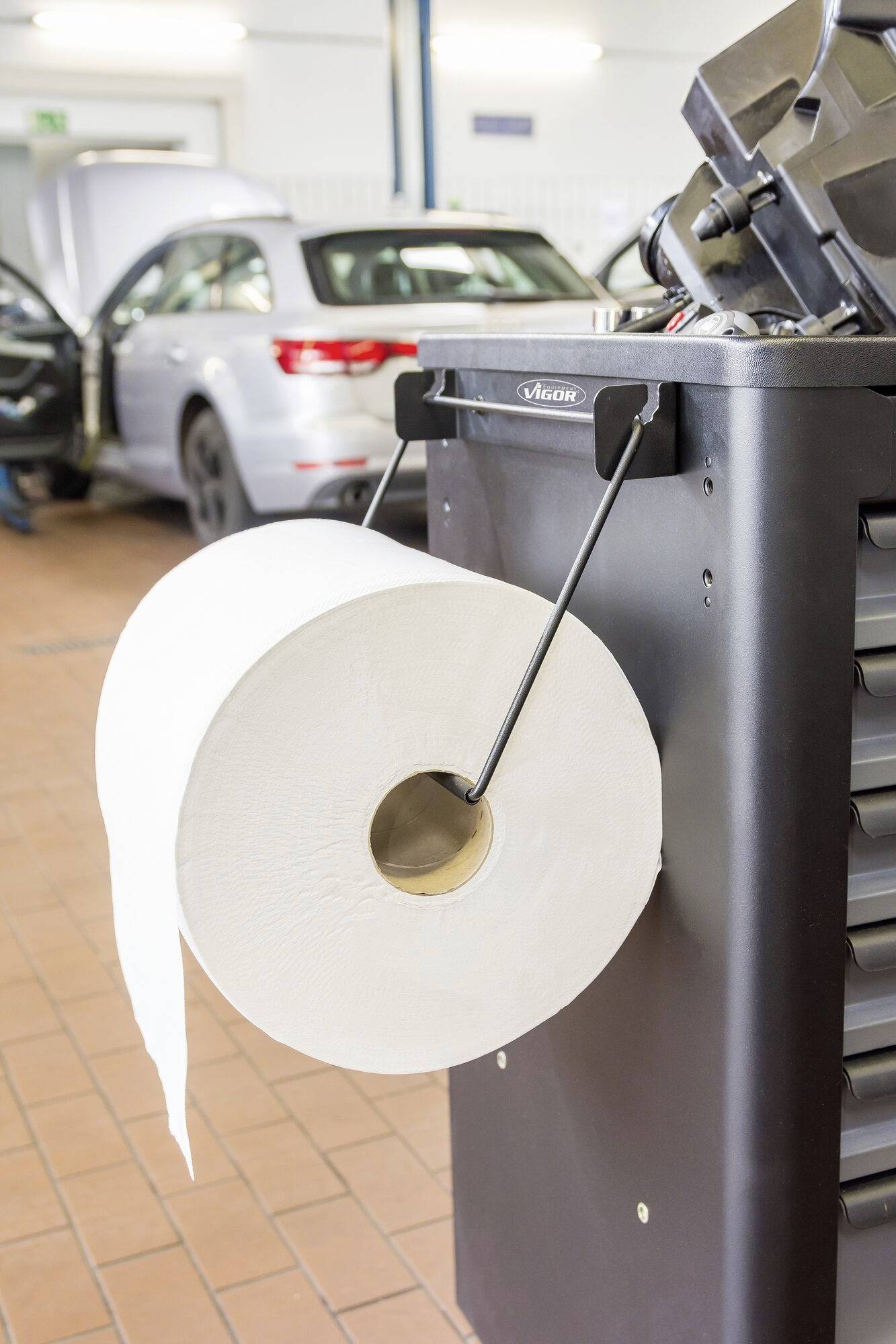 Een grote papierrol hangt aan een gereedschapswagen in een autogarage. Op de achtergrond staat een auto met de motorkap open.