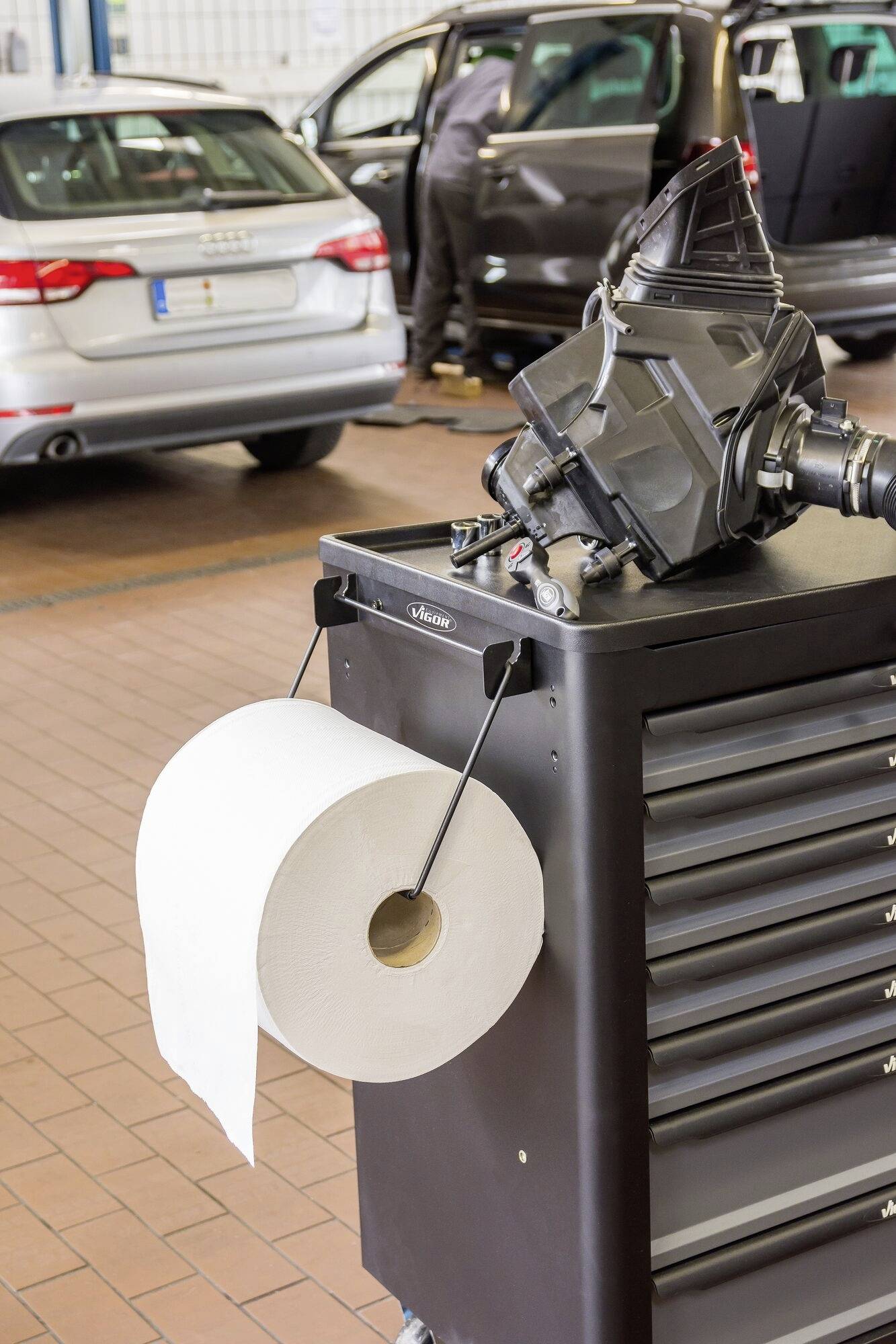 Autowerkplaats: Een werkwagen met een grote papierrol op de voorgrond staat naast een monteur die aan een auto op de achtergrond werkt.