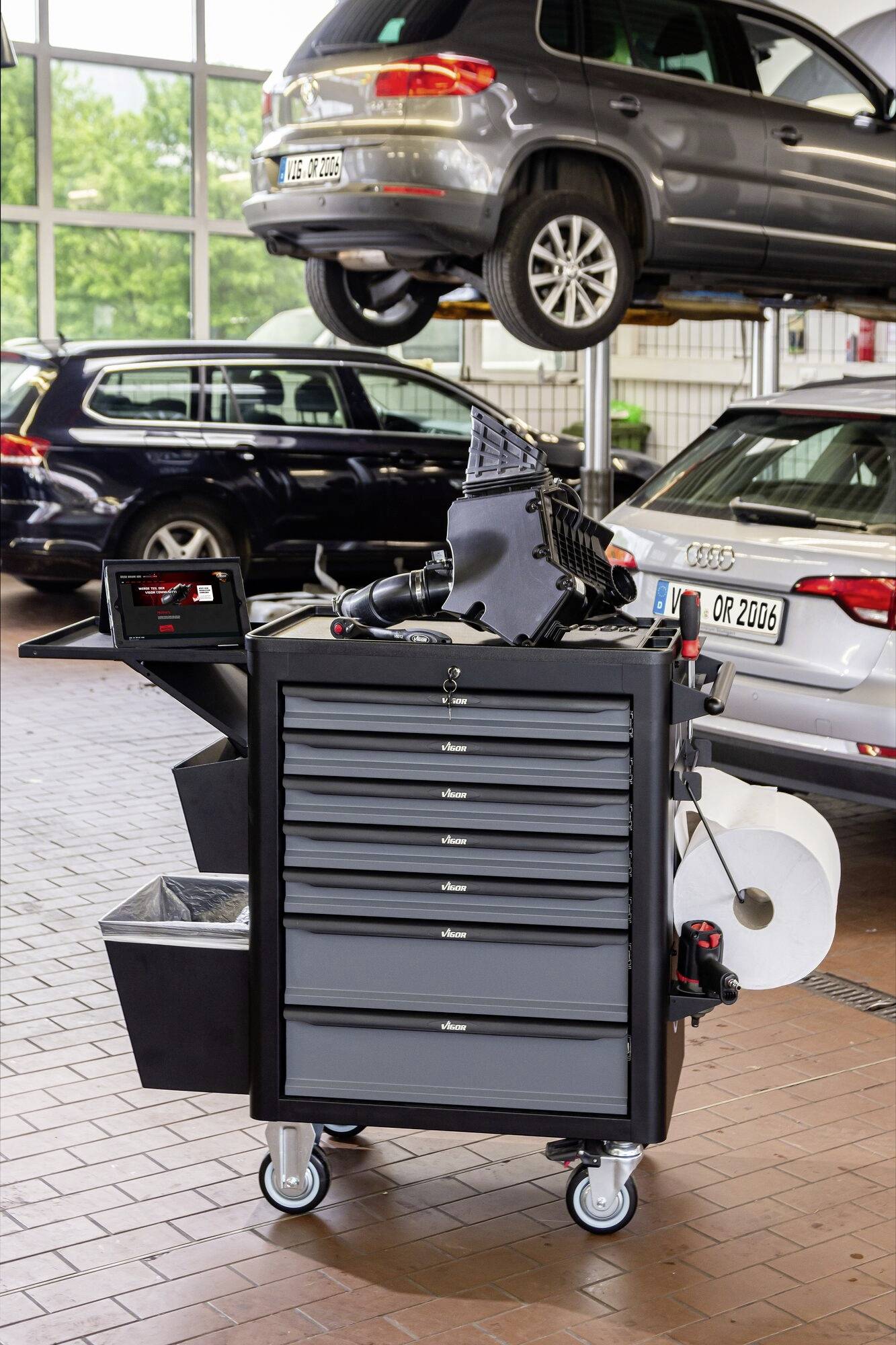 Een zwarte gereedschapswagen staat in een autowerkplaats. Op de achtergrond staat een auto op een hefbrug voor reparatiewerkzaamheden.