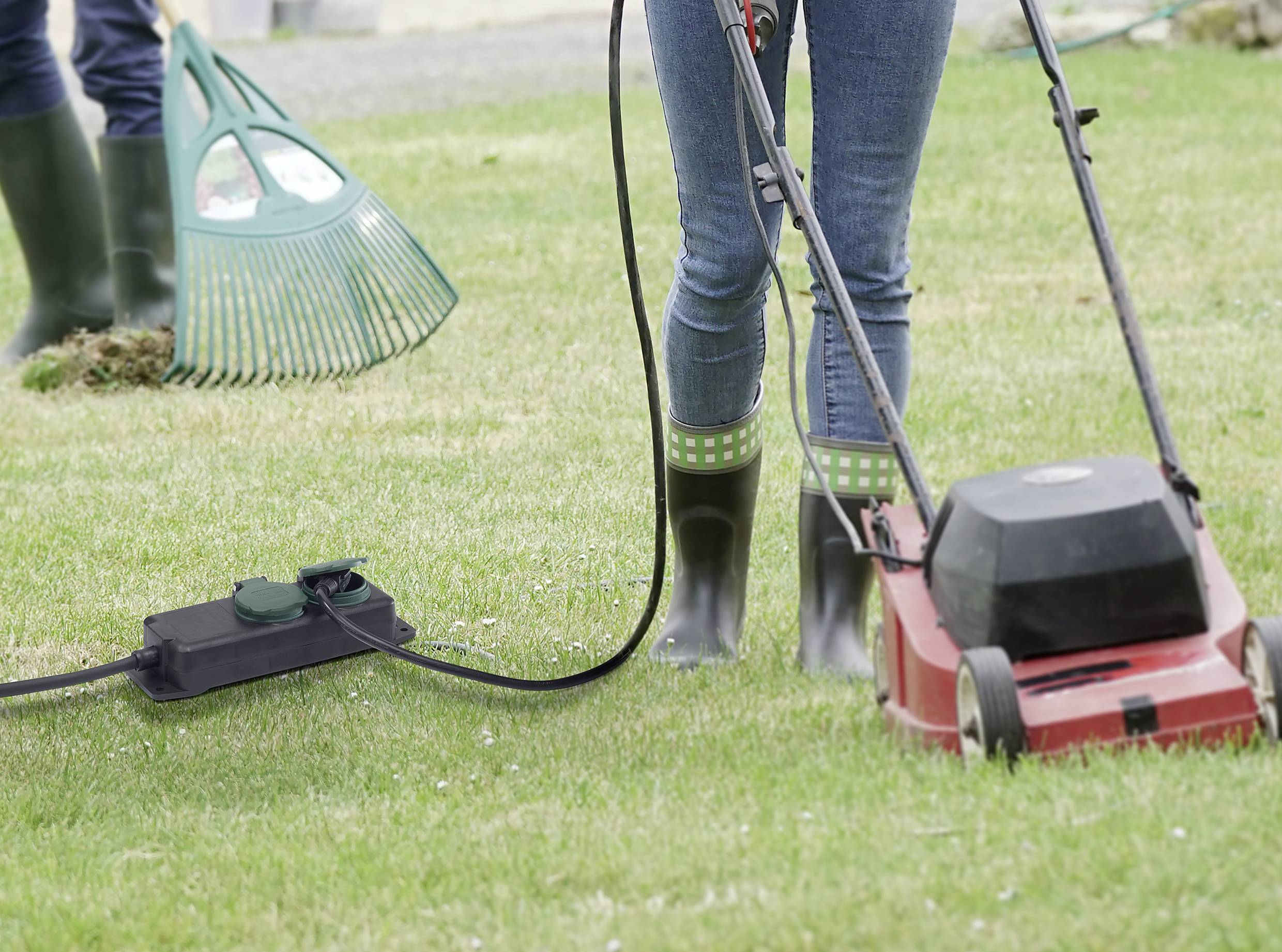 Een persoon maait het gazon met een elektrische grasmaaier. Op de achtergrond is nog een persoon te zien met een hark.