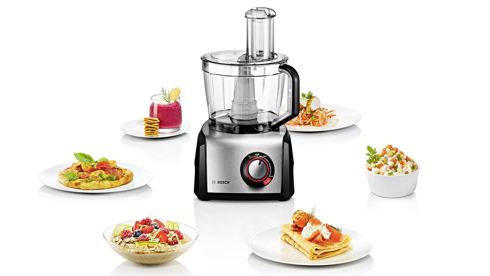 In het midden staat een keukenmachine, omringd door vijf verschillende gerechten, waaronder pannenkoeken, salade en dessert.