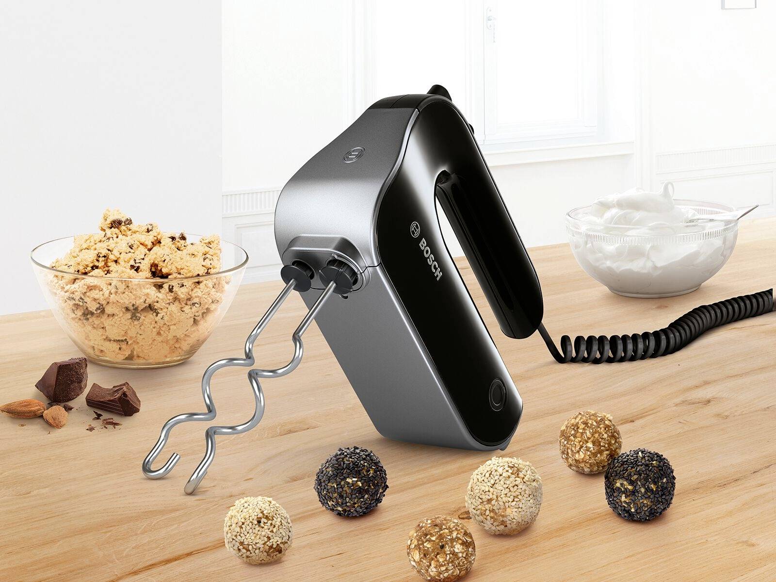 Zwarte handmixer op houten tafel, omgeven door koekjesdeeg en slagroom. Garden zijn bevestigd, kabel op de achtergrond.