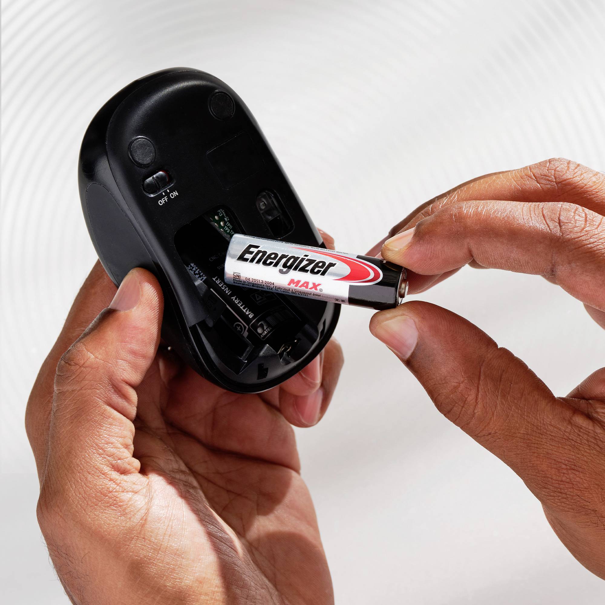 Een persoon stopt een Energizer-batterij in een draadloze computermuis. De muis staat open, en de handen houden de batterij vast.