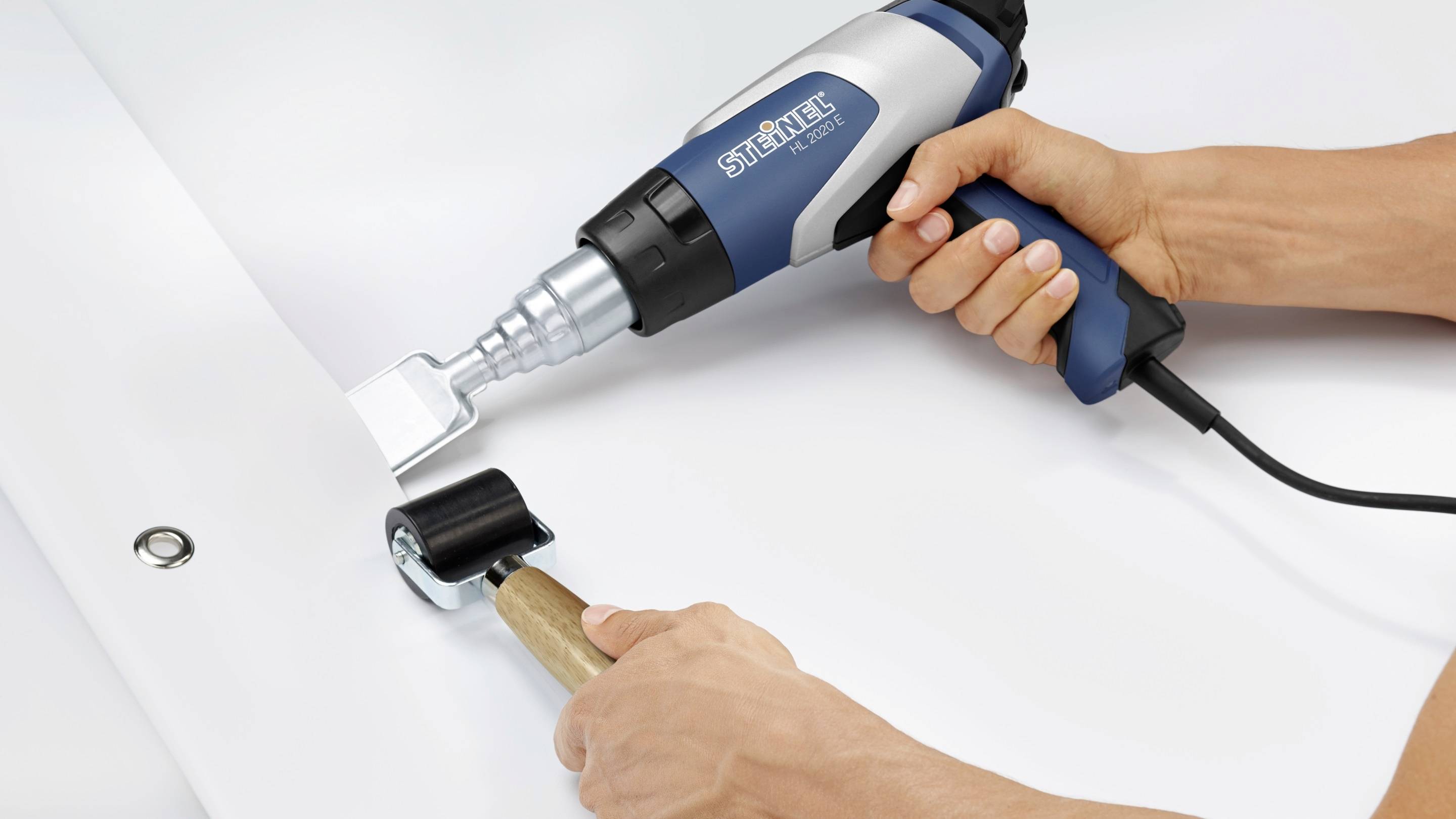 Een persoon gebruikt een blauwe heteluchtpistool om materiaal op een witte ondergrond te lijmen. Een roller effent het materiaal.