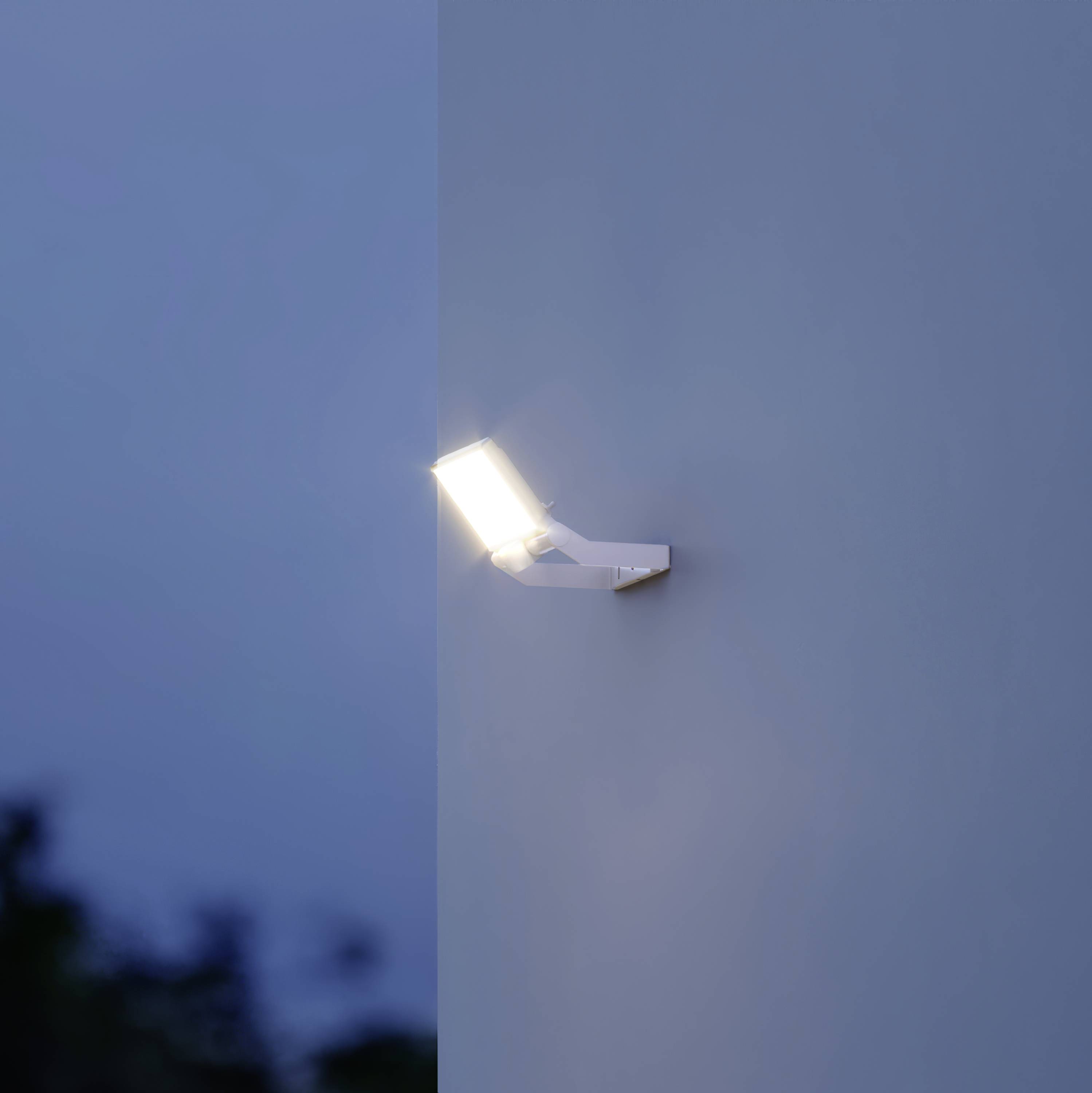 Een verlichtingsarmatuur is aan een muur bevestigd en verspreidt licht tijdens de schemering. De lucht is donkerblauw.