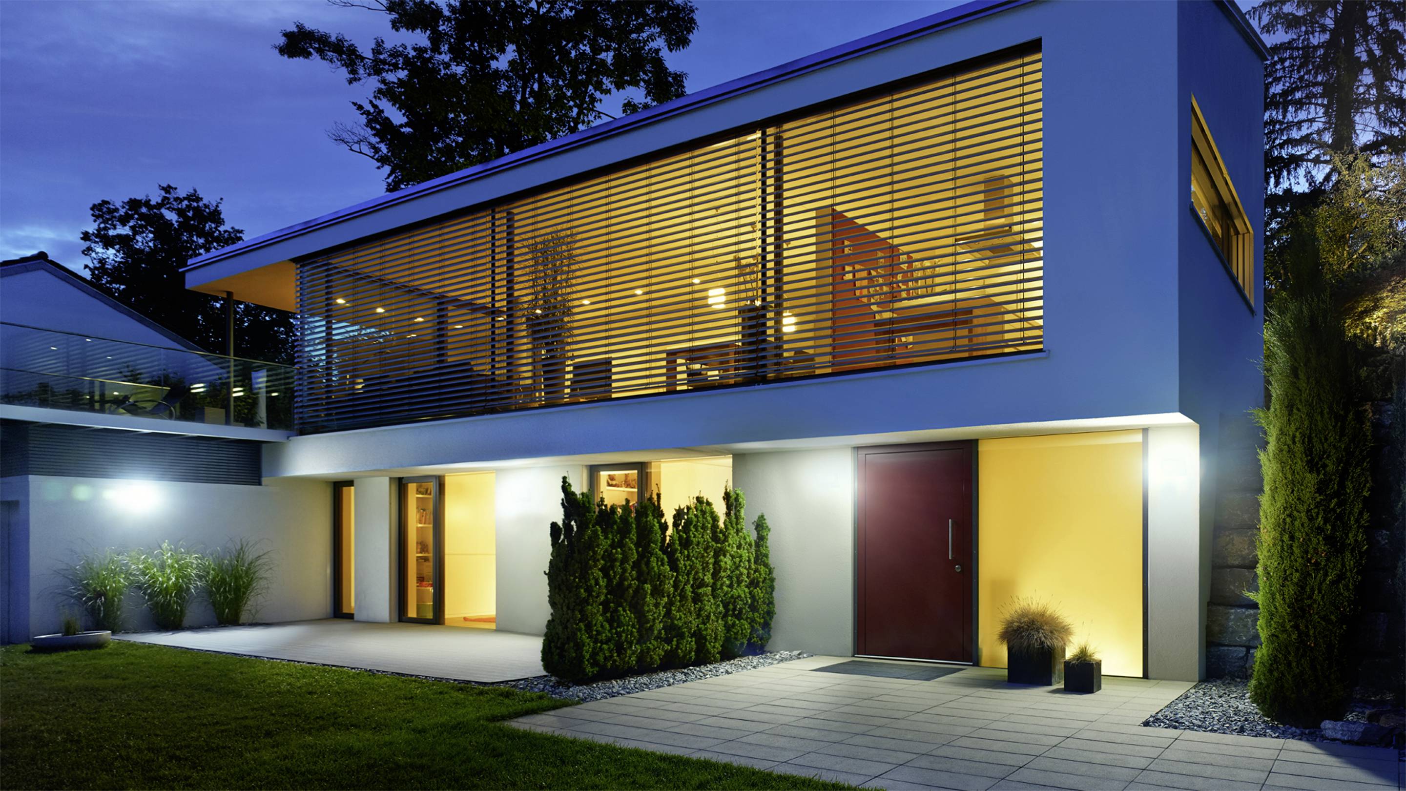 Modern tweeverdiepingshuis bij avondschemering, met grote glazen gevel op de bovenverdieping, verlichte veranda en keurig onderhouden tuin.