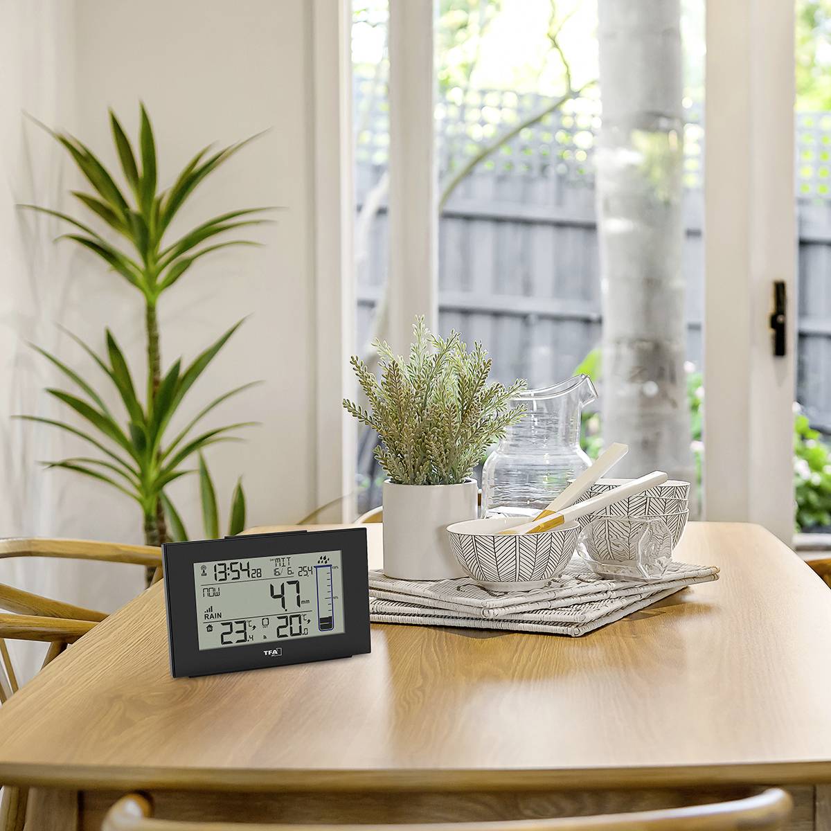 Een houten tafel met decoratieve schalen en een plant. Ernaast staat een digitale thermometer met klok. Op de achtergrond zijn grote ramen met zicht naar buiten.