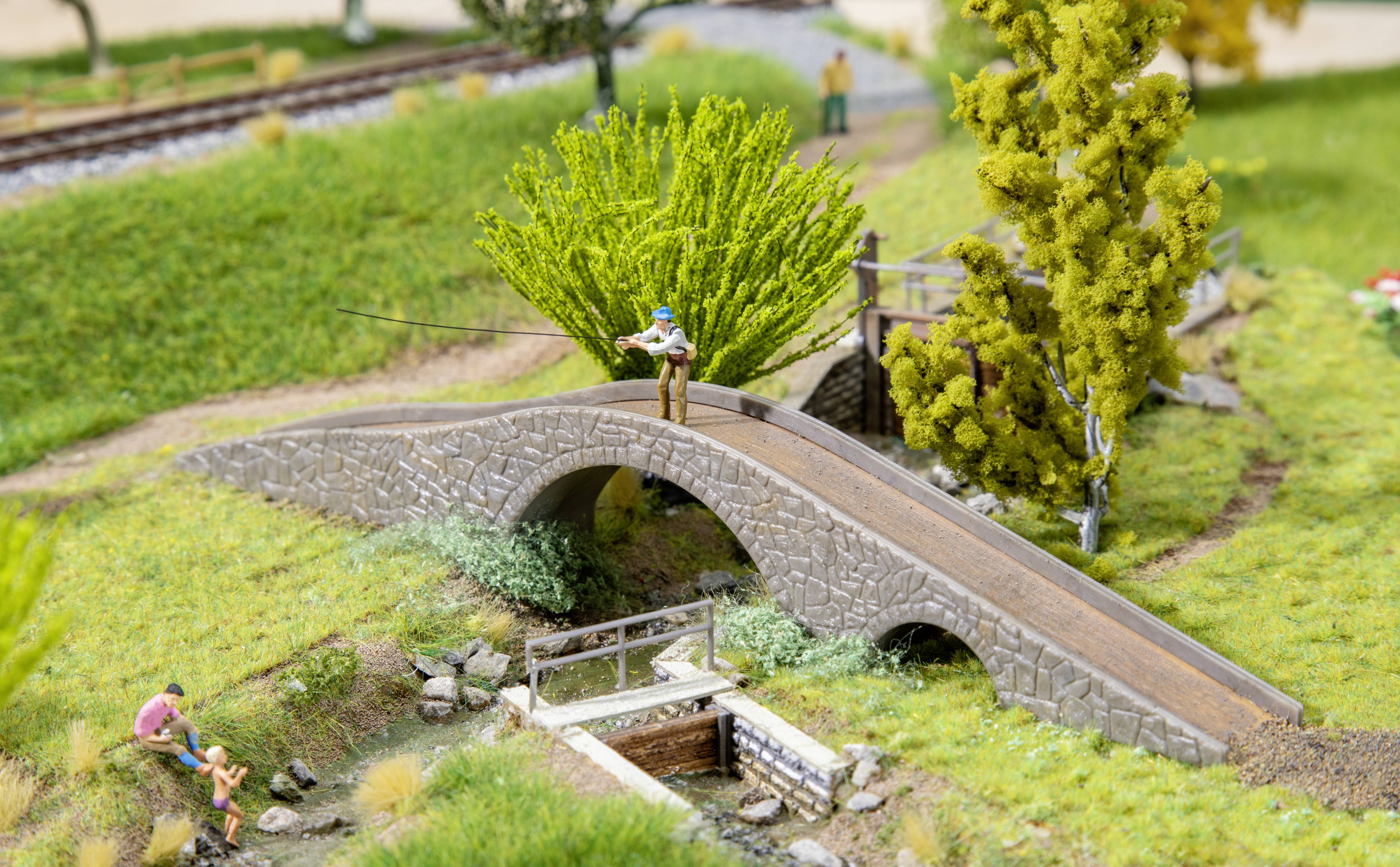 Een miniatuurmodel toont een brug over een kleine beek. Een figuur staat op de brug met een hengel, een andere zit ernaast.