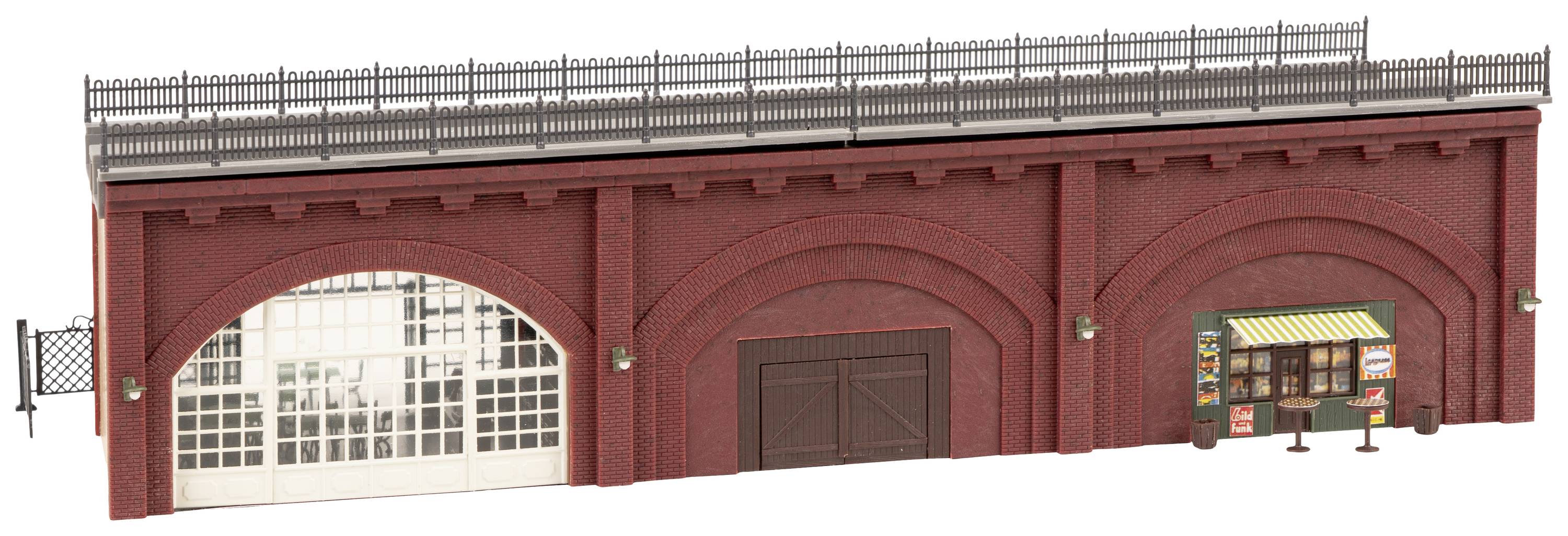 Rood bakstenen gebouw met bogen, een kleine winkel en een hek bovenaan. Een poort en etalages zijn zichtbaar.