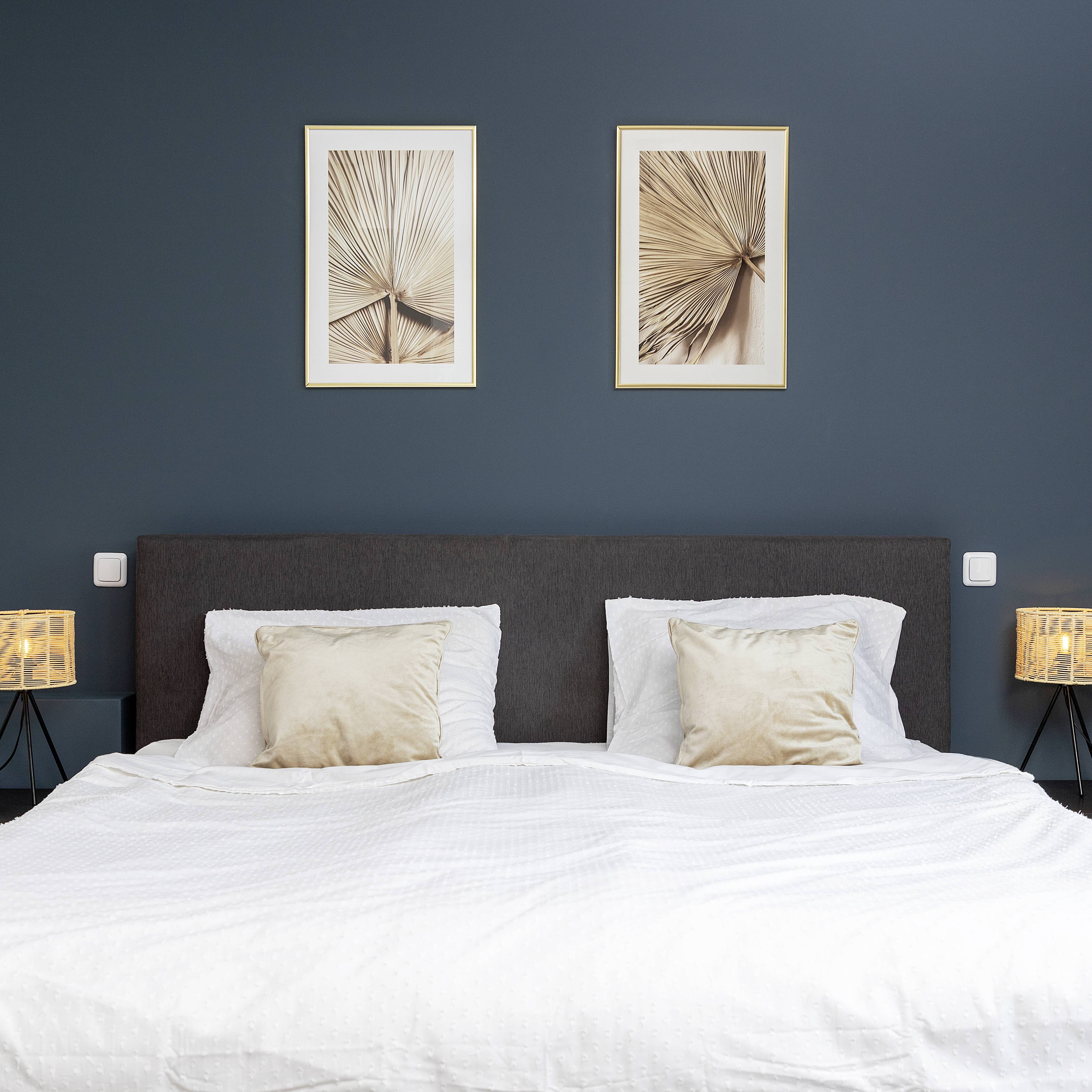 Een modern slaapkamer met een tweepersoonsbed, twee beige kussens, een blauw-grijze muur en twee abstracte kunstwerken erboven.