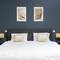 Een modern slaapkamer met een tweepersoonsbed, twee beige kussens, een blauw-grijze muur en twee abstracte kunstwerken erboven.