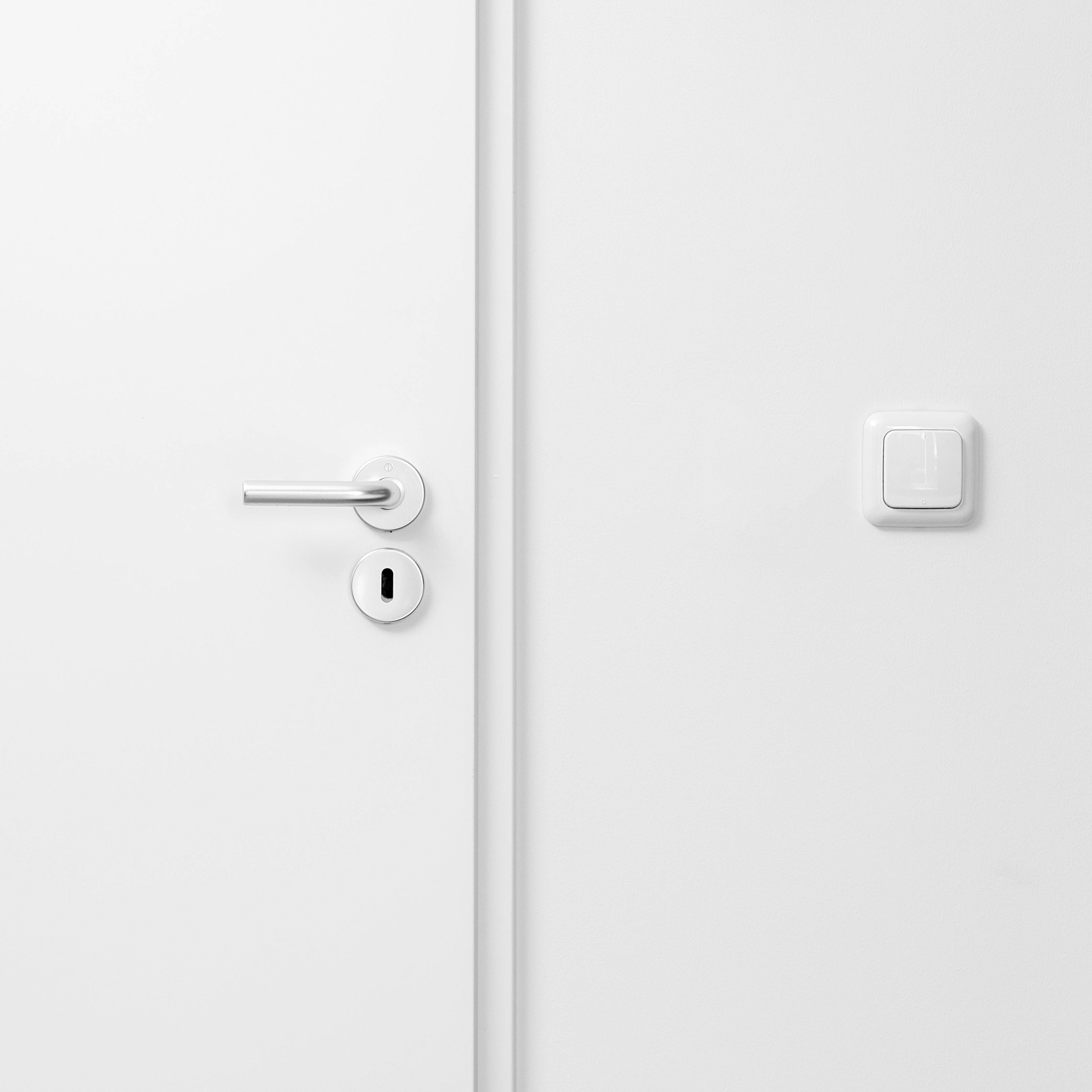 Witte deur met zilveren deurklink en sleutelgat aan de linkerkant; lichtschakelaar rechts naast de deur. Minimalistisch design.