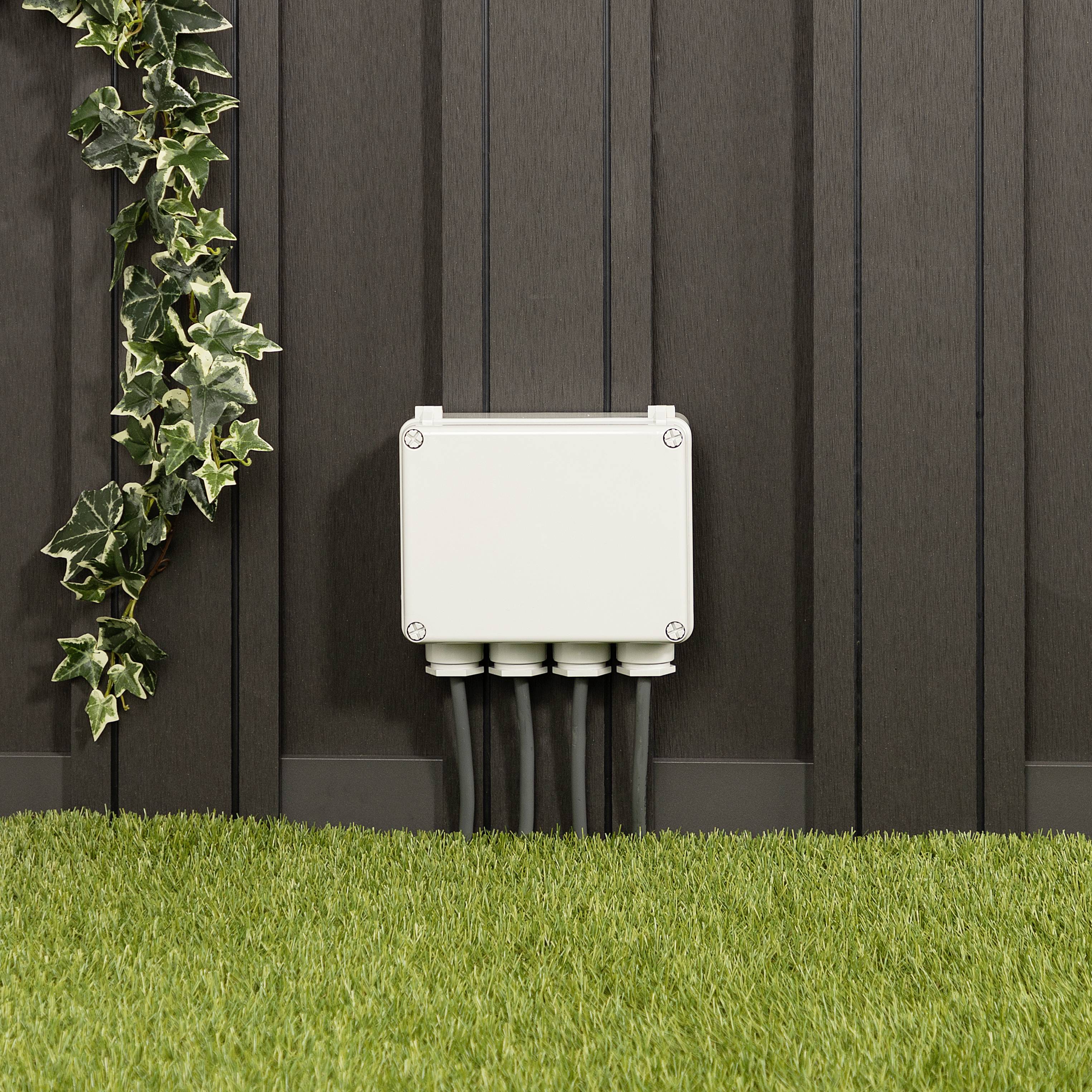 Een elektrische verdeelkast is gemonteerd aan een houten muur. Kabels lopen naar beneden. Links slingeren groene klimopranken.