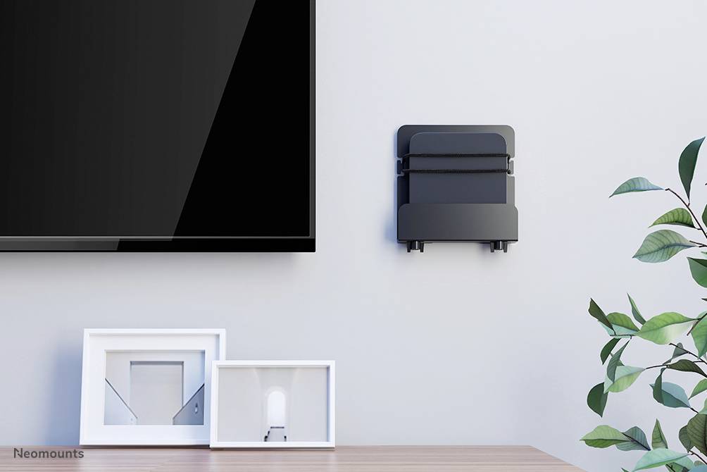 Flatscreen tv aan de muur naast een decoratief wandrek met twee fotolijsten, waarvan een leeg en een met kunst. Plant rechts in beeld.
