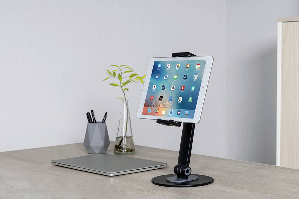 Een tablet op een zwarte standaard, geplaatst op een bureau naast een gesloten laptop, een vaas met planten en een pennenbak.