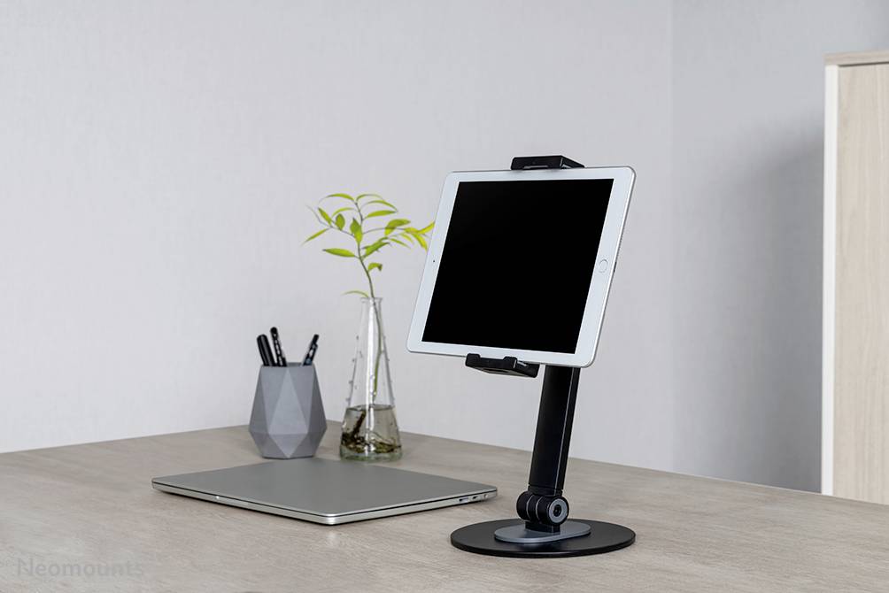 Een tablet staat in een verstelbare houder op een bureau naast een gesloten laptop en een pennenhouder.