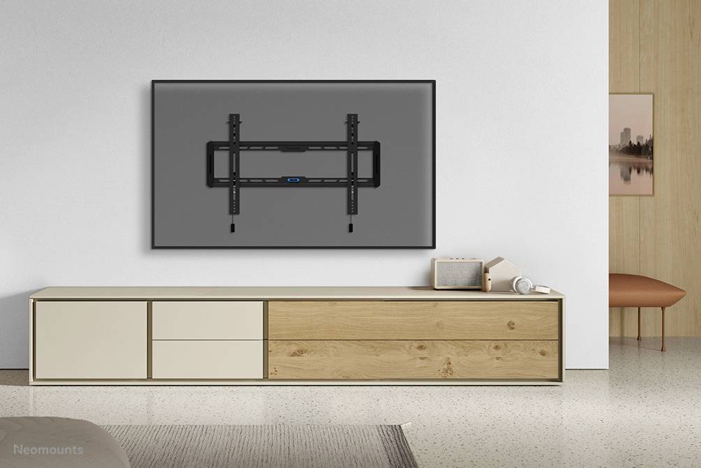 Wandbevestiging voor tv gemonteerd aan een witte muur, voor een minimalistische wit-beige tv-console met houten details. Links staat een kleine luidspreker.