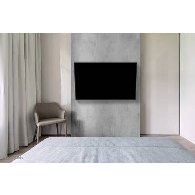 Eenvoudige slaapkamer met grijze muur waarop een flatscreen-tv is gemonteerd. Links staat een fauteuil naast lange gordijnen.
