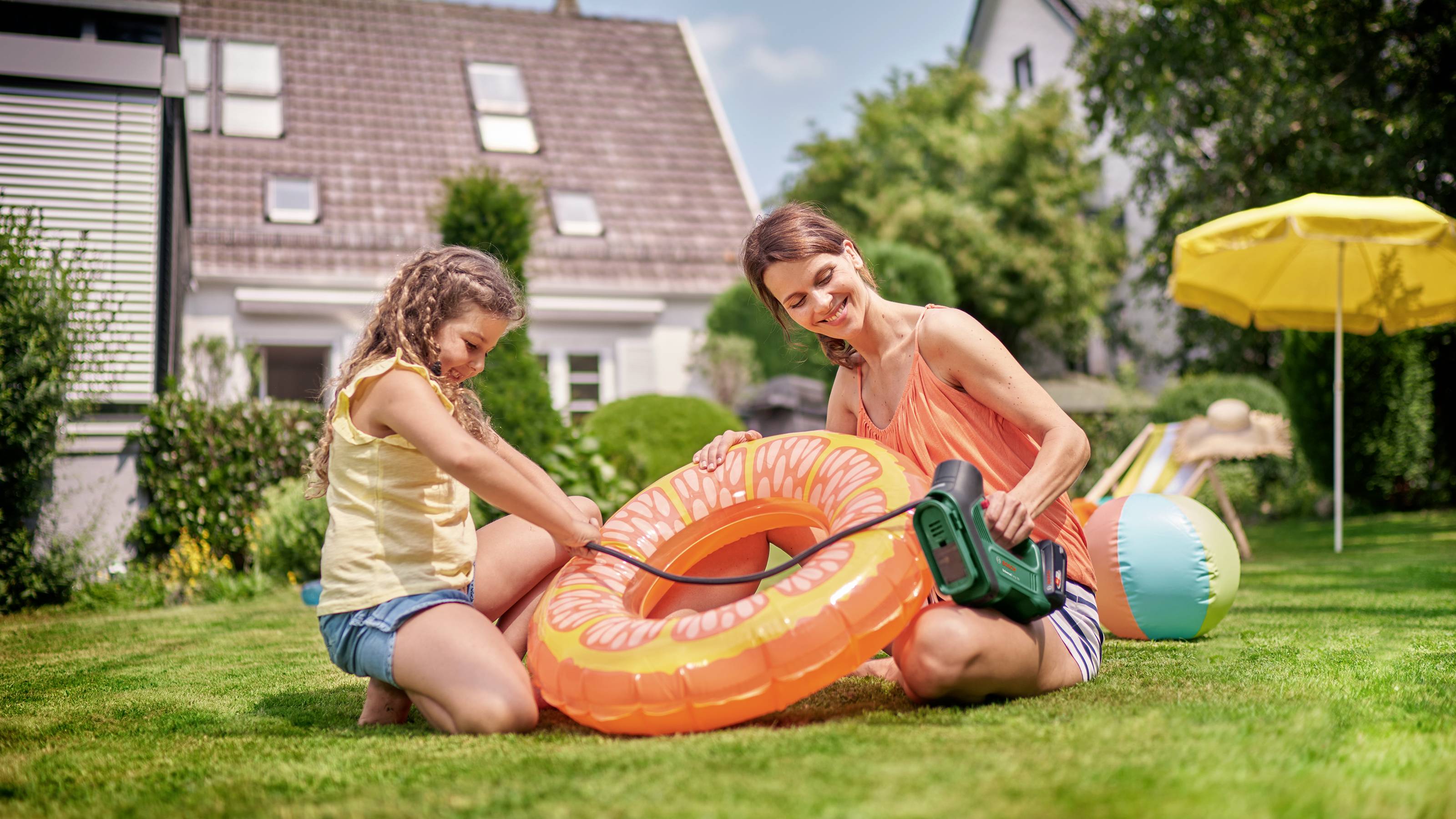 Een vrouw en een kind zitten in de tuin en pompen een oranje zwemband op met een elektrische pomp. Achtergrond: zon, huizen.