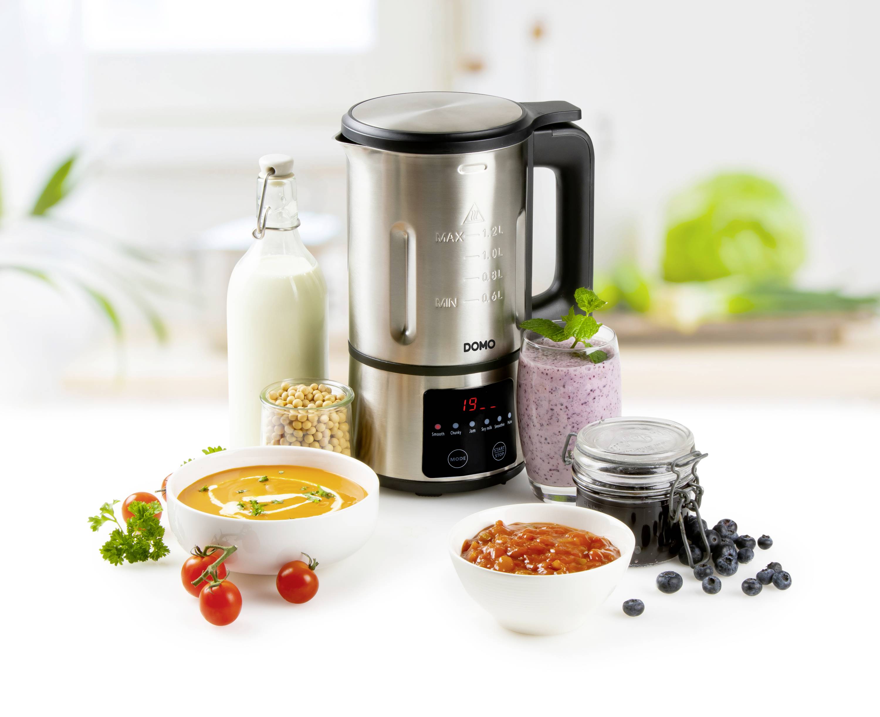 Een mixer staat op een tafel, omringd door een fles melk, een kom soep, een kom saus, een glas met bessen en kikkererwten.
