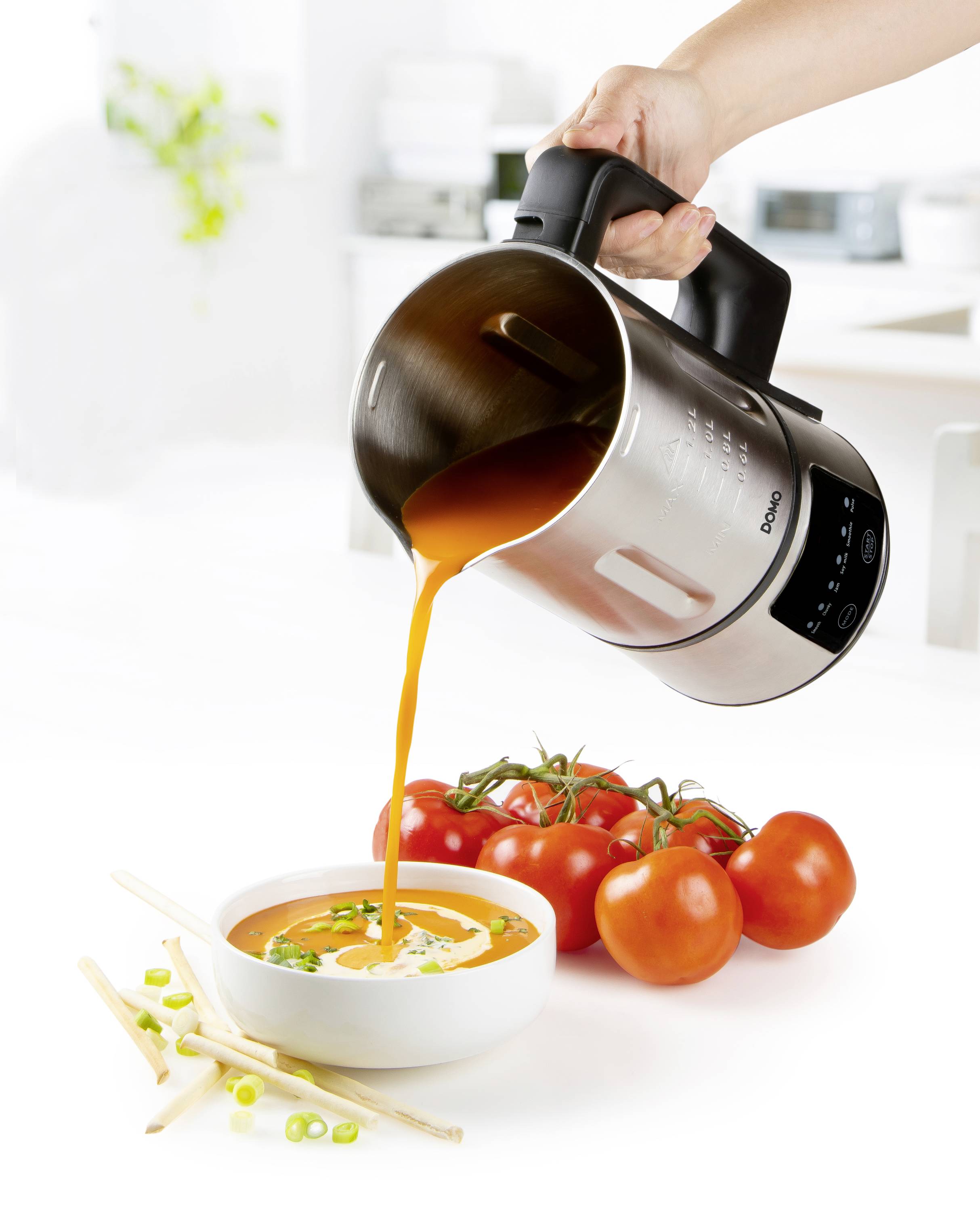 Een hand giet soep uit een zilveren staande blender in een witte kom. Ernaast liggen verse tomaten en lente-uitjes.