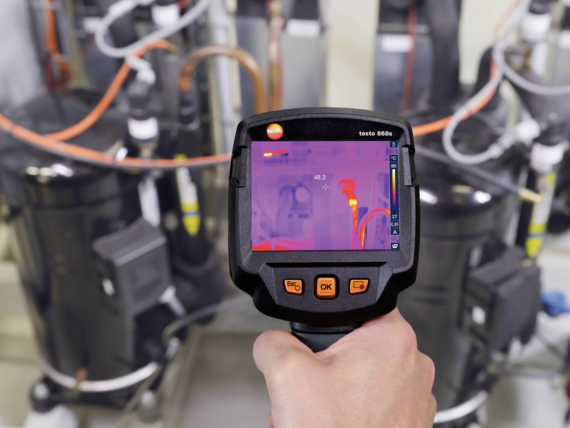 Een persoon houdt een thermische camera die een temperatuurmeting weergeeft. Achtergrond toont technische apparatuur.