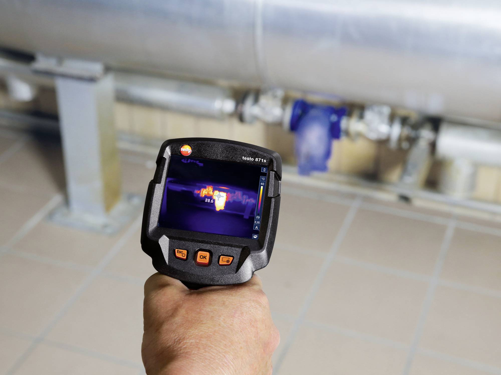Een persoon houdt een warmtebeeldcamera vast die een verwarmingsbuis inspecteert. Het display toont temperatuurverschillen langs de buis.