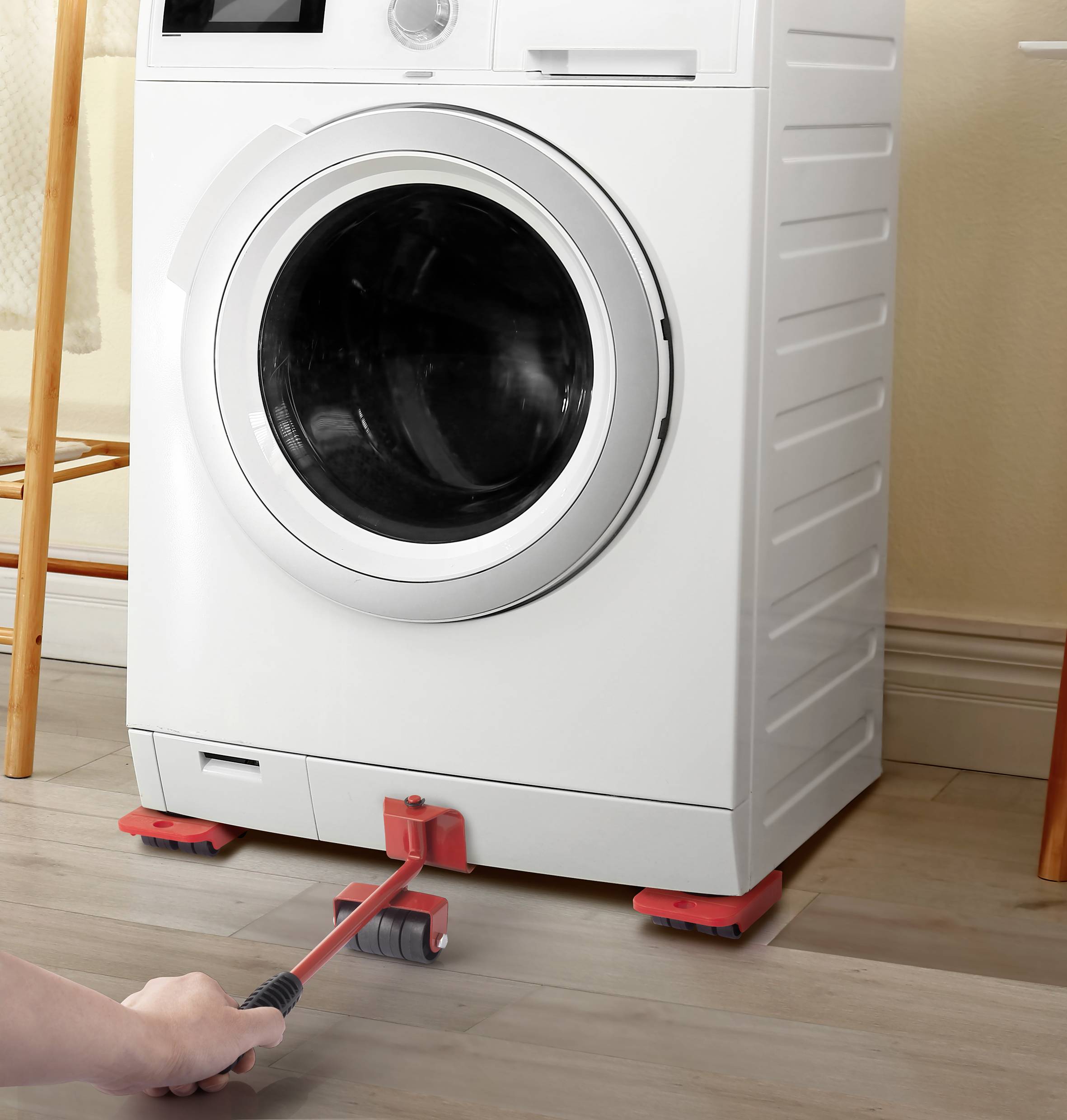 Een wasmachine op een houten vloer. Een hand gebruikt een hefboomgereedschap met wieltjes eronder om de machine te verplaatsen.
