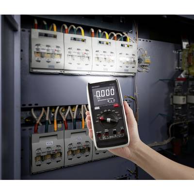 Een persoon houdt een digitale multimeter voor een open schakelkast met meerdere bekabeling en zekeringen.
