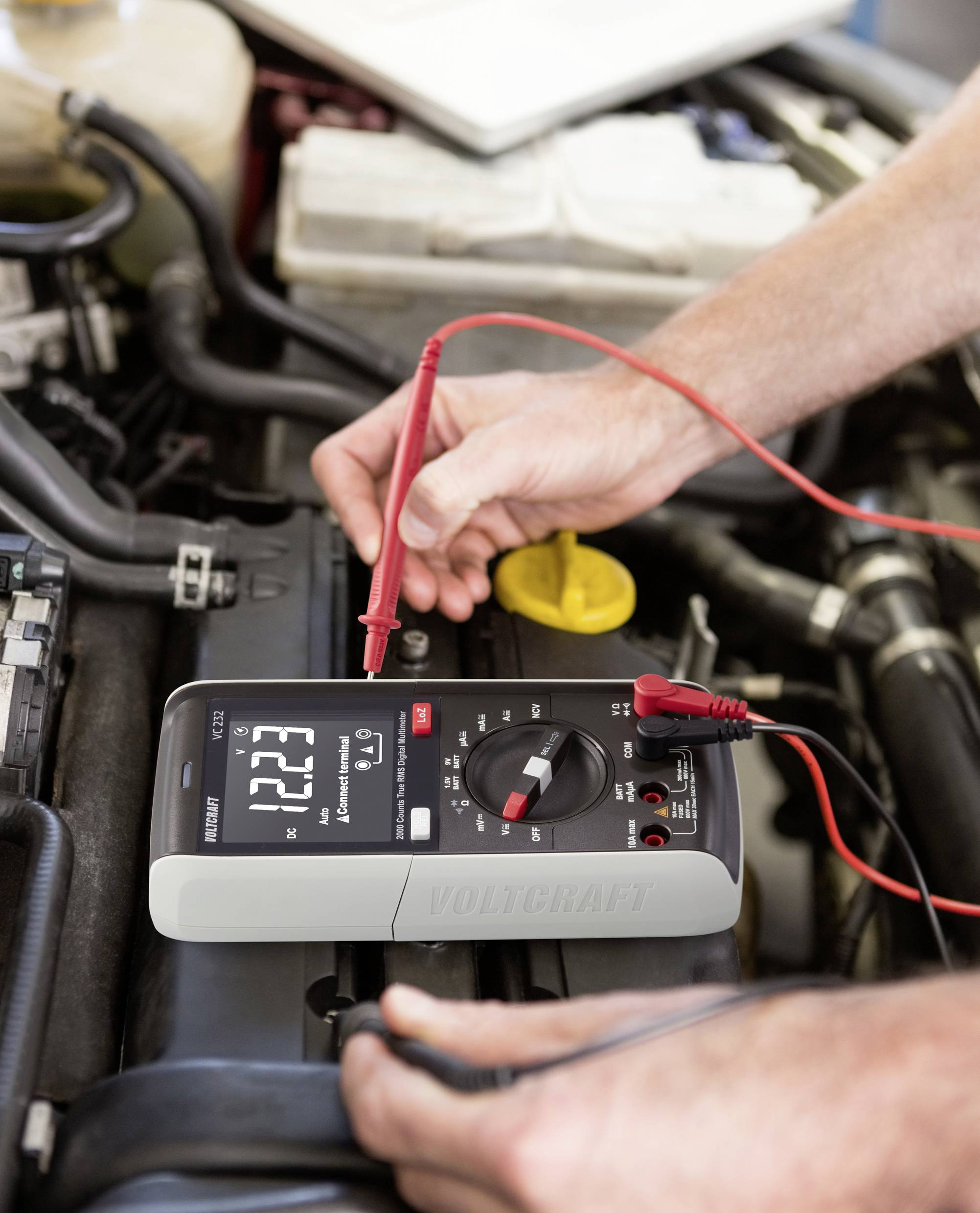 Persoon meet de accuspanning van een auto met een digitaal multimeter dat 12,23 volt aangeeft. Kabels zijn aangesloten.