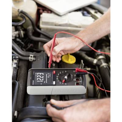 Persoon meet de accuspanning van een auto met een digitaal multimeter dat 12,23 volt aangeeft. Kabels zijn aangesloten.