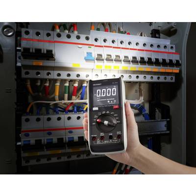 Een hand houdt een digitale multimeter vast die een spanning van 0,000 aangeeft, voor een geopende elektrakast met meerdere kabels.