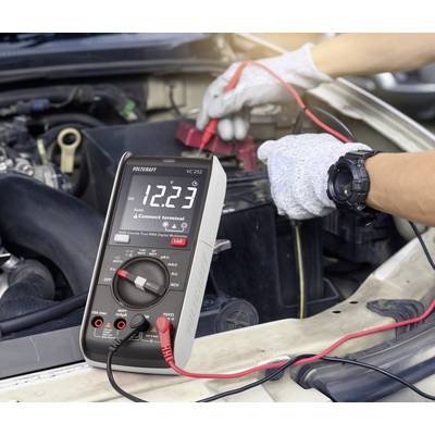 Een multimeter geeft 12,23 volt aan terwijl deze is aangesloten op een autobatterij in de motorruimte. Een persoon met handschoenen houdt de klemmen vast.