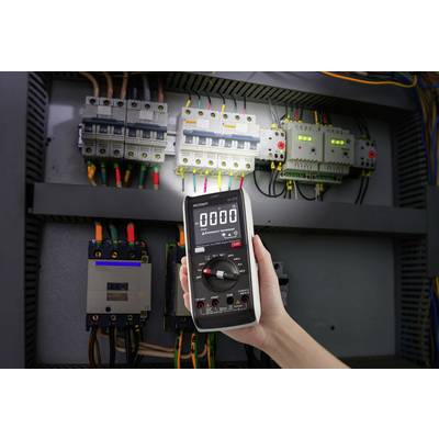 Een hand houdt een digitale multimeter vast die nul aanwijst, voor een geopende elektrische schakelkast met zekeringen en kabels.