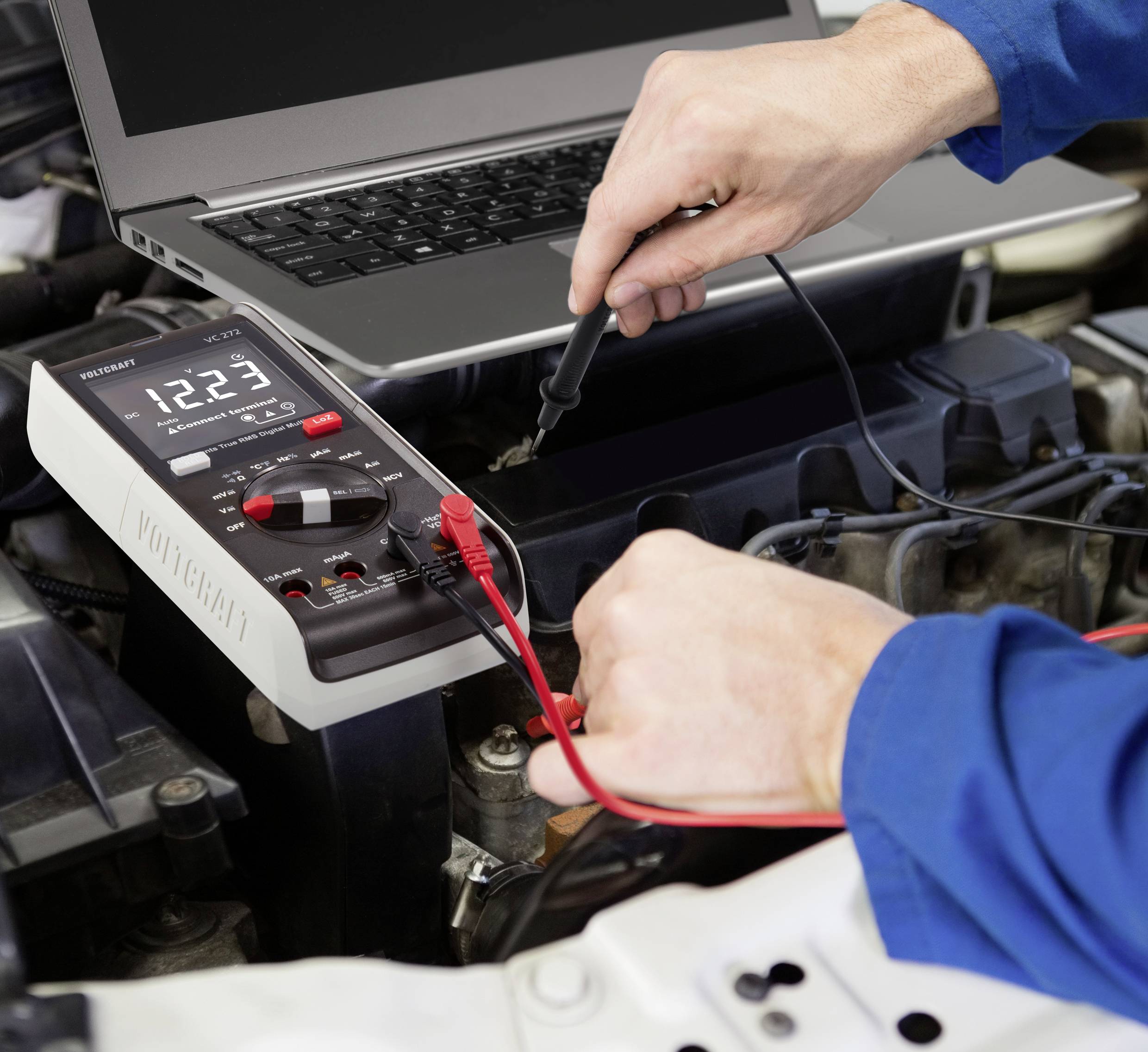 'Een persoon test de autobatterij met een multimeter, terwijl naast hem een laptop geopend is.'