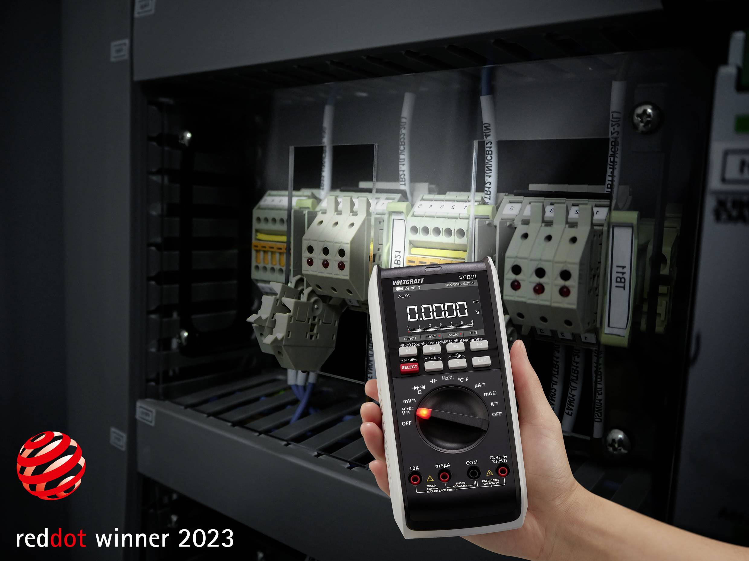 Een hand houdt een digitale multimeter voor een schakelkast met elektrische componenten, 'reddot winner 2023' in de hoek.