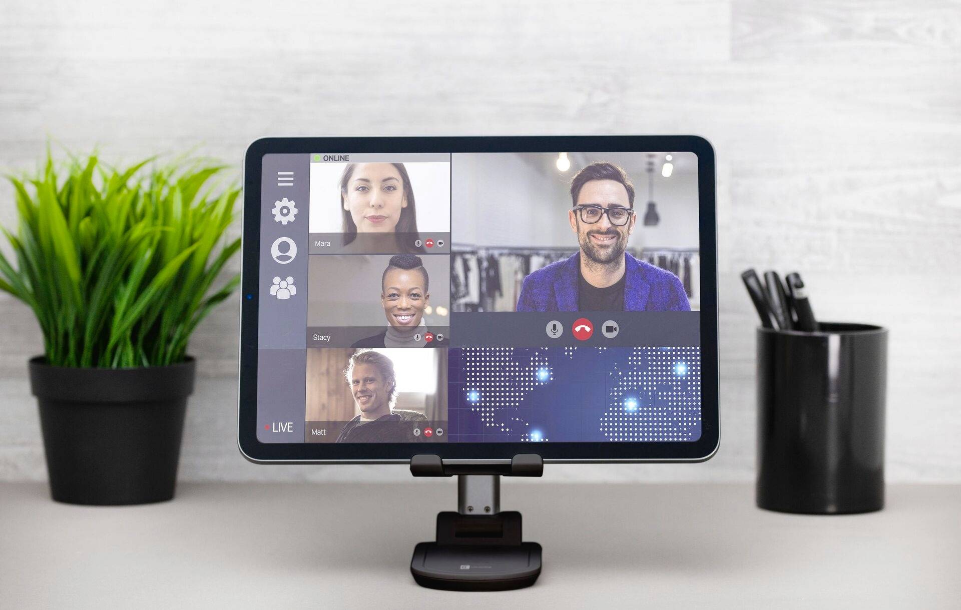 Een tablet toont een videoconferentiegesprek met vier deelnemers. Links ernaast een plant, rechts een pennenhouder op een bureau.
