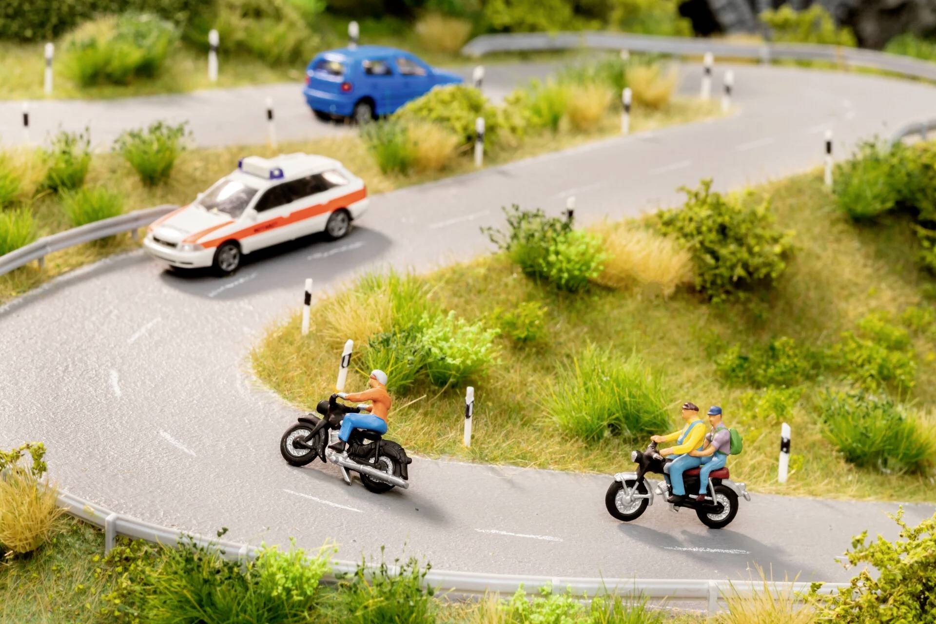 Een miniatuurlandschap toont een kronkelende weg met twee motoren en een auto. Er zijn mensen te zien op de motoren.