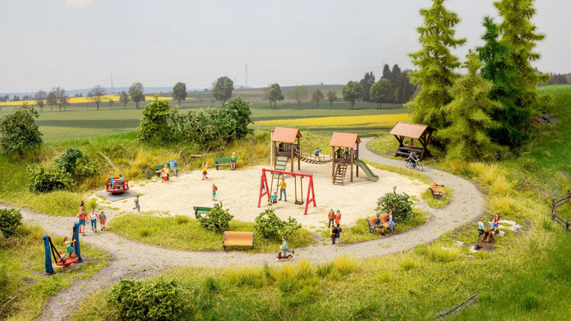 Een speelplaats in een landelijke omgeving met schommels, glijbanen en kinderen die aan het spelen zijn. Omringd door bomen en weilanden.
