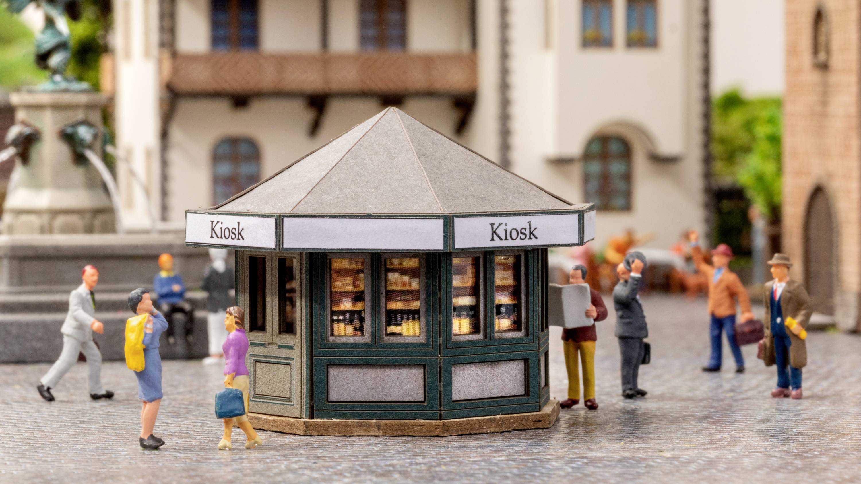 Een kleine, achthoekige kiosk op een stadsplein met mensen die voorbijlopen en blijven staan. Gebouwen op de achtergrond.