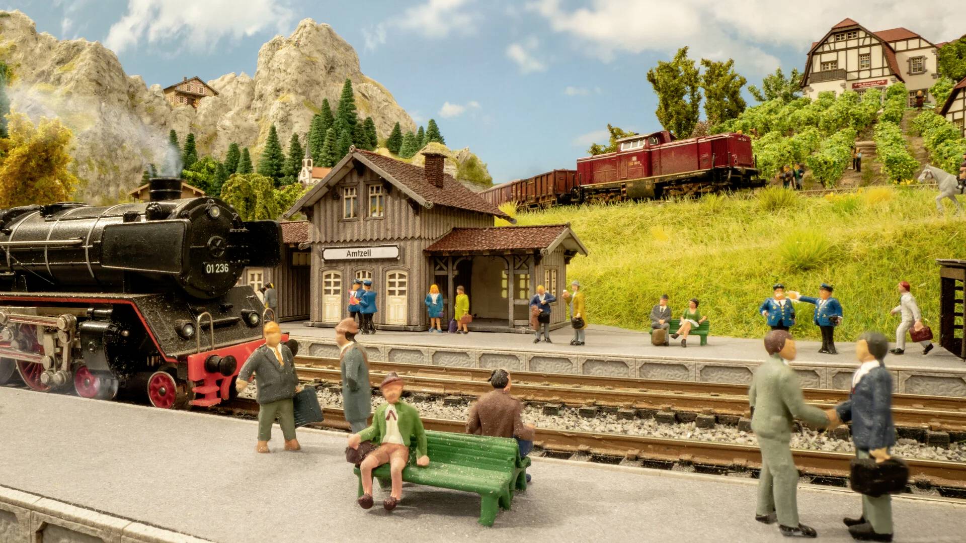 Een miniatuurspoorscène met een trein bij het station. Figuurtjes staan of zitten, omringd door een landschap met bergen en bomen. Gebouwen op de achtergrond.