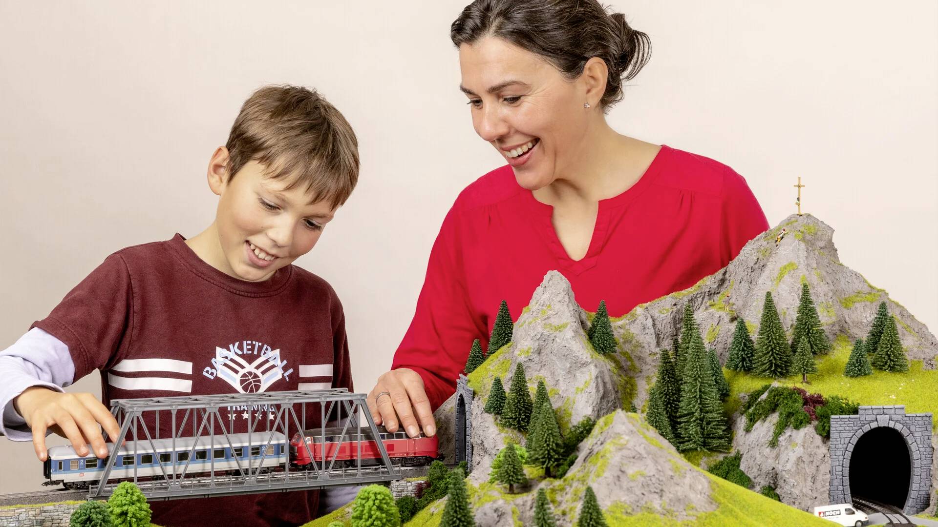 Een vrouw en een jongen spelen samen met een modelspoorbaan in een landschap met bergen, een brug en een tunnel.