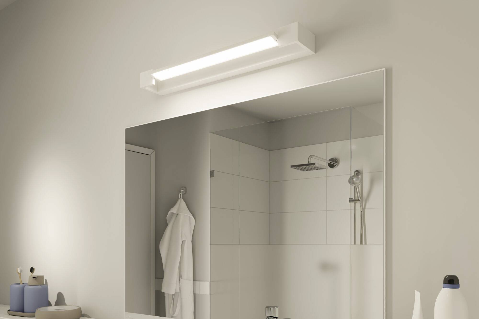Een badkamer met een grote spiegel, daarboven een verlichte lamp. Links zijn toiletartikelen, op de achtergrond een douche.