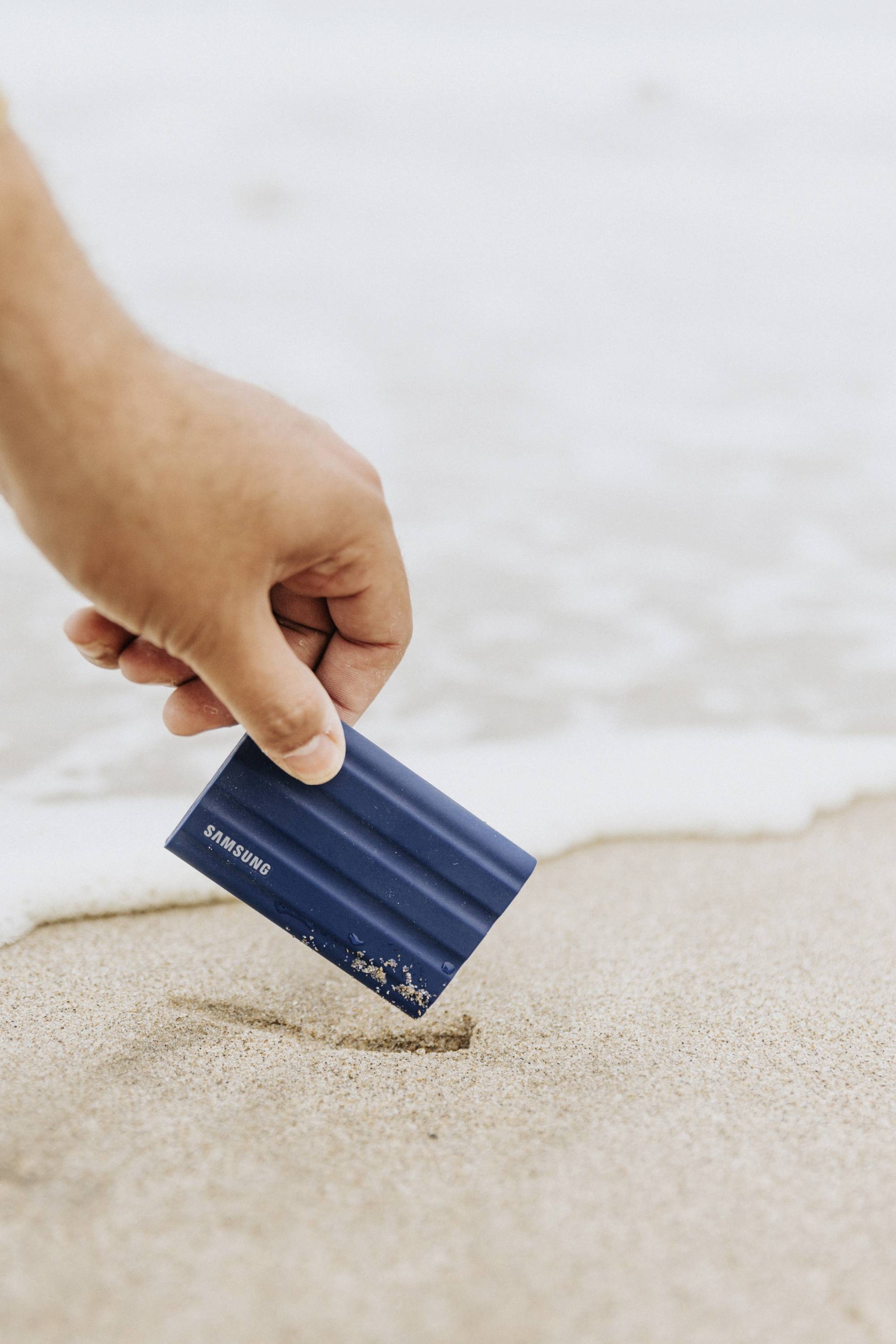 Een hand tilt een klein blauw apparaat op uit het zand op een strand. Op de achtergrond is de zachte branding zichtbaar.