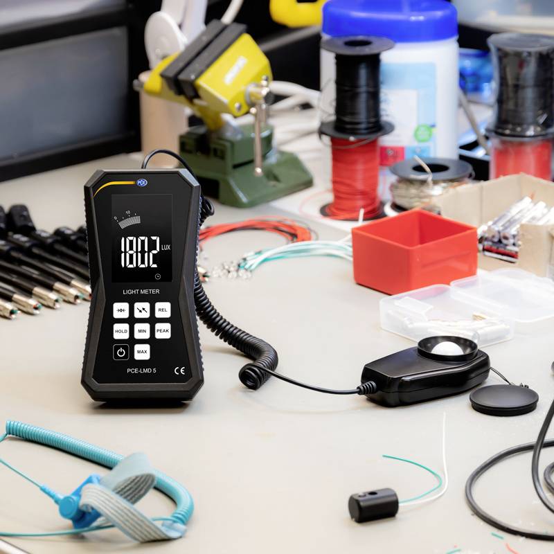 Een tafel toont een apparaat voor lichtmeting bij 180,2 Lux, evenals gereedschappen en kabels voor elektronische werkzaamheden op de achtergrond.