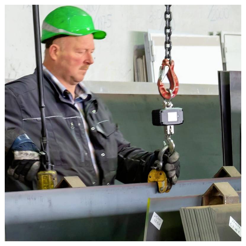 Fabrieksarbeider in een groene helm gebruikt een kraanhaak en hefklem om een grote metalen balk te verplaatsen.