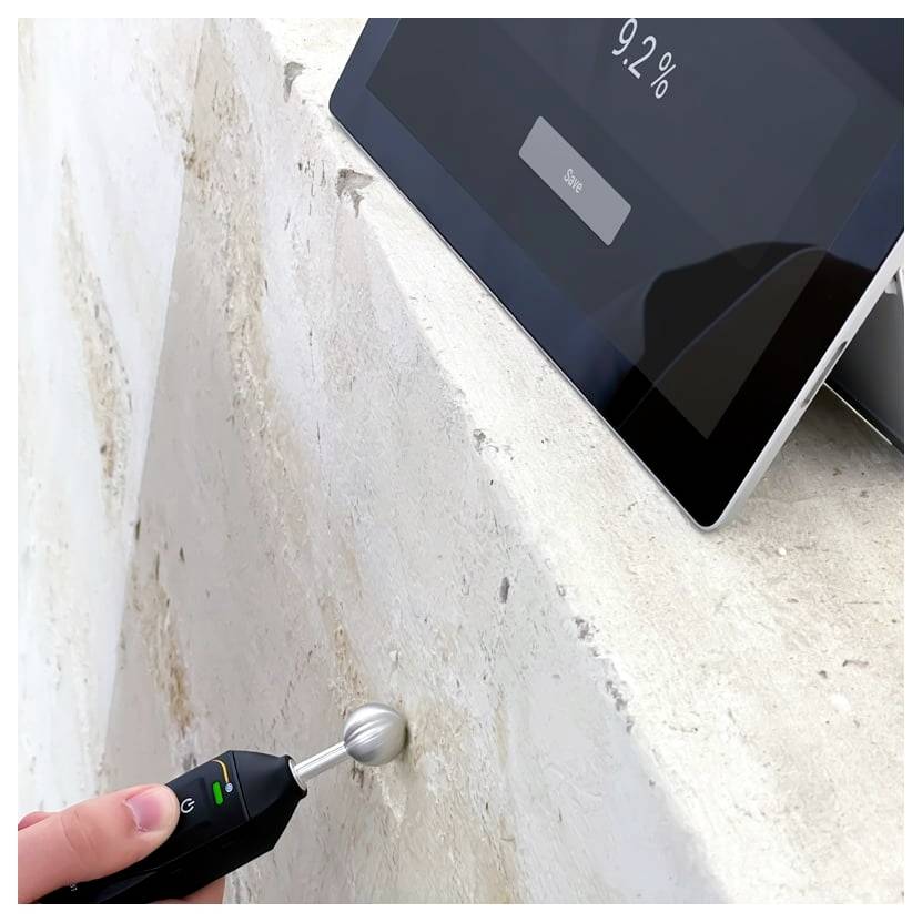 Handheld vochtmeter test aan een muur terwijl een tablet 92% luchtvochtigheid aangeeft, wat duidt op vochtigheid of waterschade.