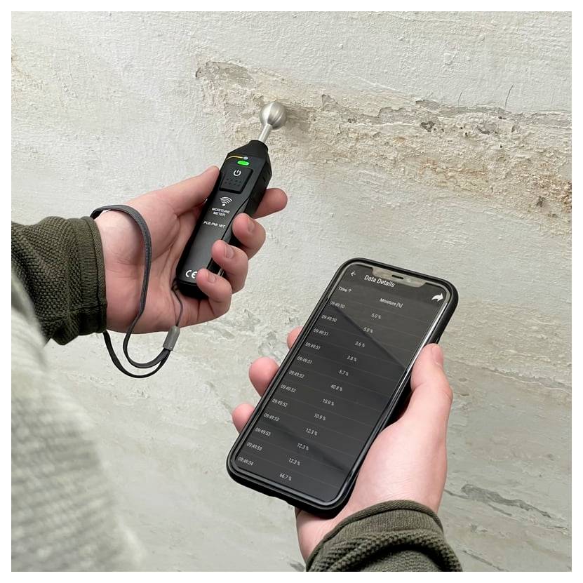 Een persoon houdt een vochtmeter tegen een vochtige, afbladderend muur terwijl hij vochtmetingen op een smartphone-app bekijkt.