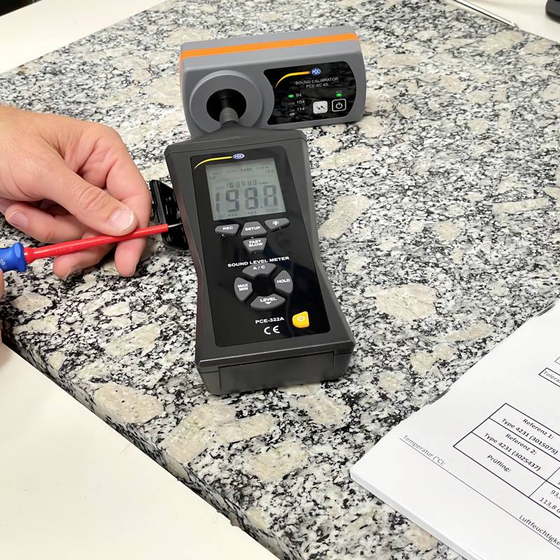 Een hand houdt een spanningsmeter vast die 198 V aangeeft op een stenen vloer, naast een zwart en grijs meetapparaat.